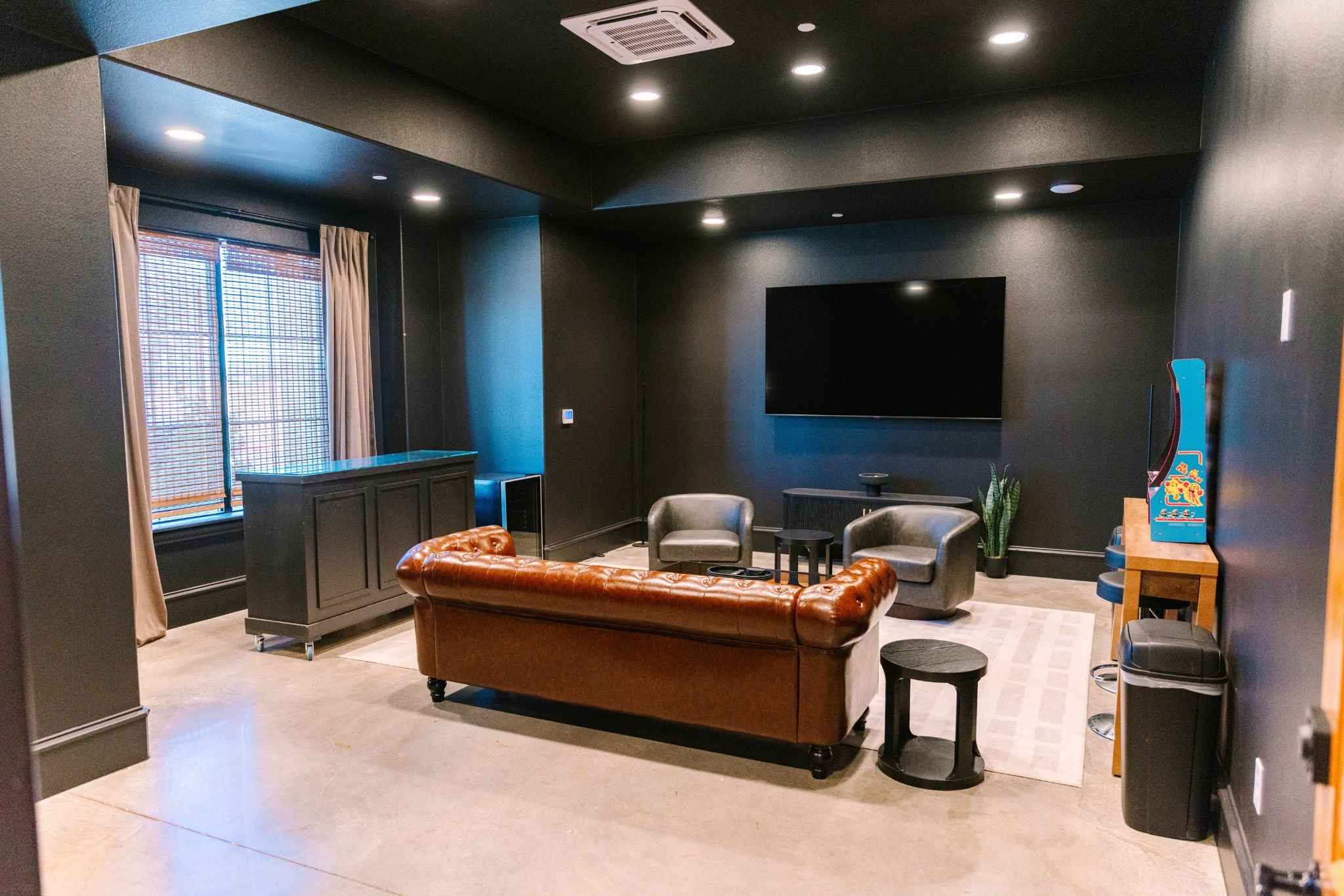 A sophisticated and masculine groom's room at Adelphos Cellars featuring a brown leather Chesterfield sofa, grey armchairs, and dark black walls. The room is equipped with a large wall-mounted TV, a retro arcade machine, and a bar area, all set on polished concrete floors with warm recessed lighting