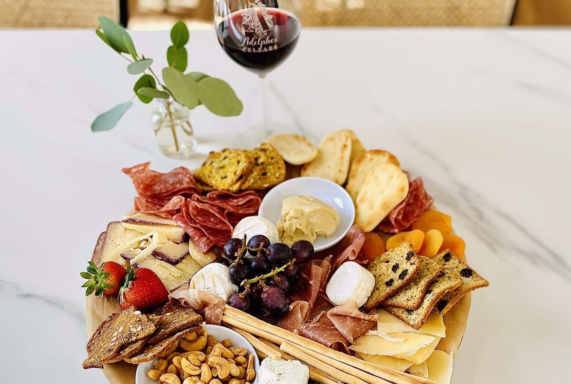A small charcuterie board with meats, cheese, fruit, and breads paired with red wine, sitting on white granite countertop inside Adelphos Cellars' tasting room